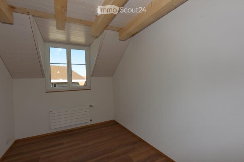 Empty attic room with wooden floor, white walls, exposed wooden beams, and a single window offering a view of the outside.