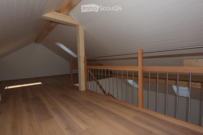 An empty attic space with wooden flooring, exposed wooden beams, and a metal railing leading to a staircase. The ceiling has skylight windows allowing natural light to enter.
