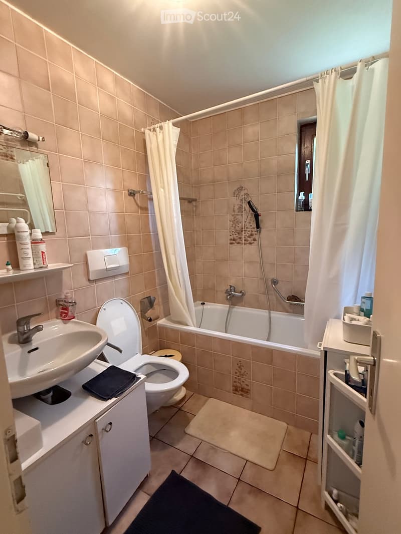 Bathroom with beige tiles, a sink, toilet, bathtub, mirror, and a small window. Includes a shower curtain, a showerhead, and a cabinet with toiletries.
