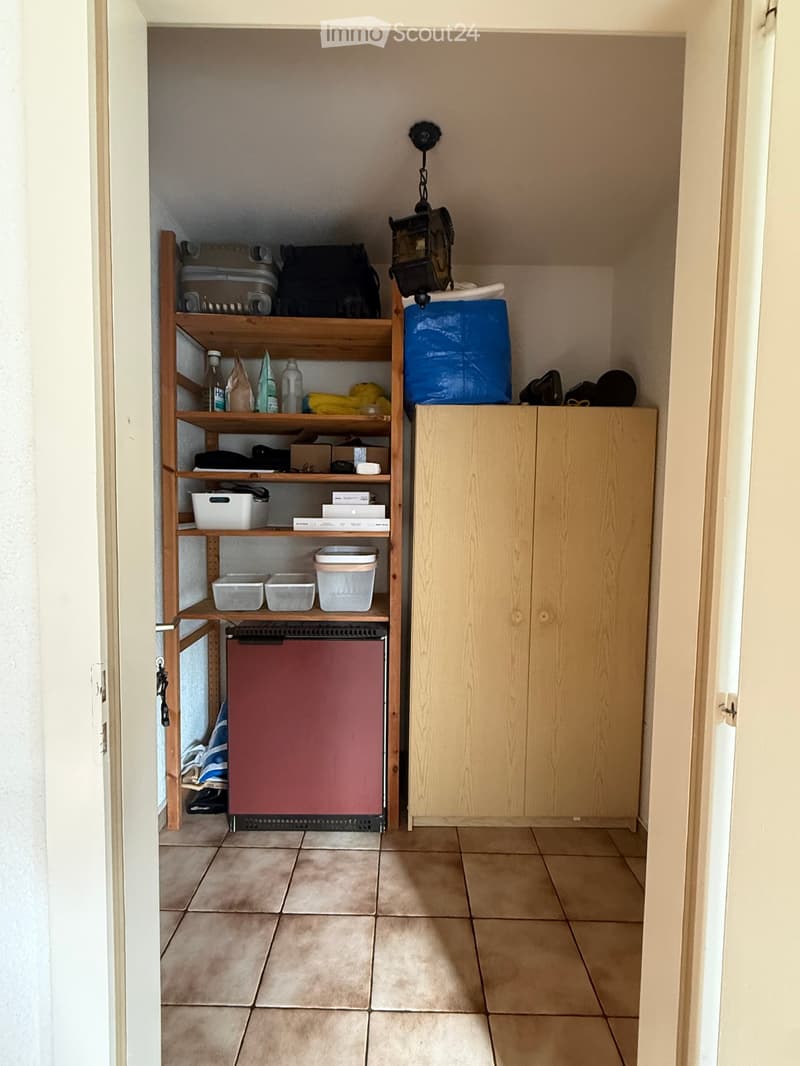 A small storage room with tiled flooring, a wooden shelf unit filled with various items, a closed wooden cabinet, and a red refrigerator. The room also contains storage bins, a bottle, and a backpack on the shelf.