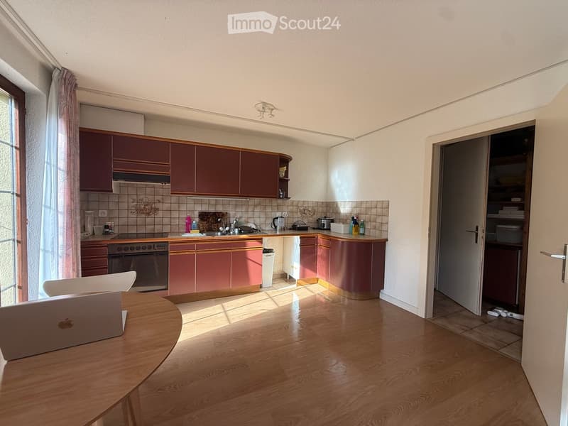 A modern kitchen with wooden flooring, red cabinets, a round wooden table with a laptop, and a chair. The kitchen features an oven, sink, and various appliances.