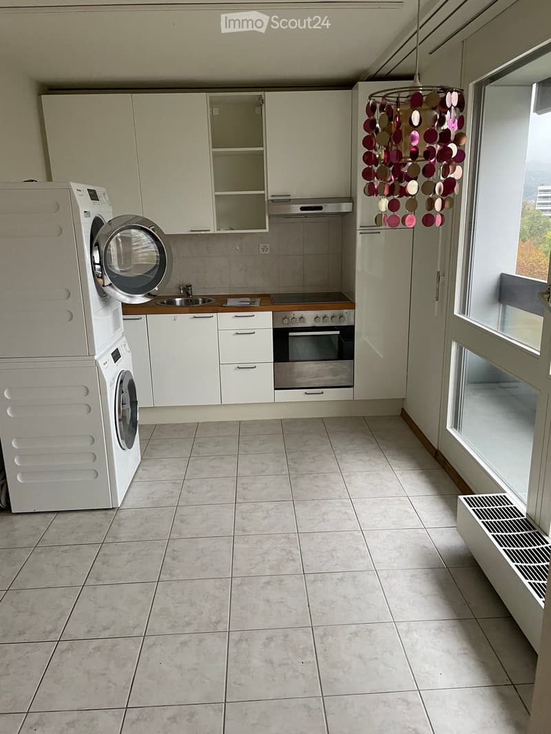 Modern kitchen with white cabinets, a washing machine, an oven, and a sink, featuring a large window and a decorative hanging light fixture.
