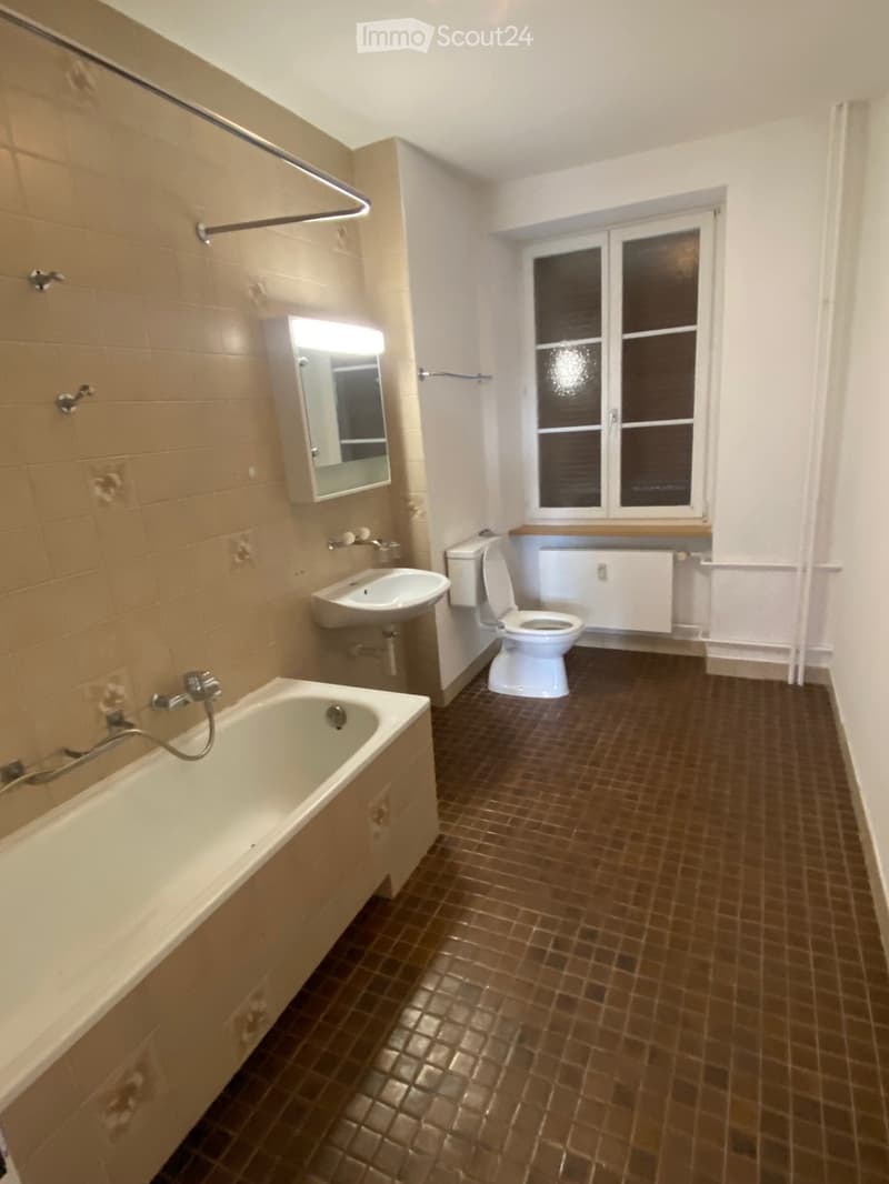 Bathroom with a bathtub, toilet, sink, mirror, and radiator. Brown tiled floor and beige tiled walls.