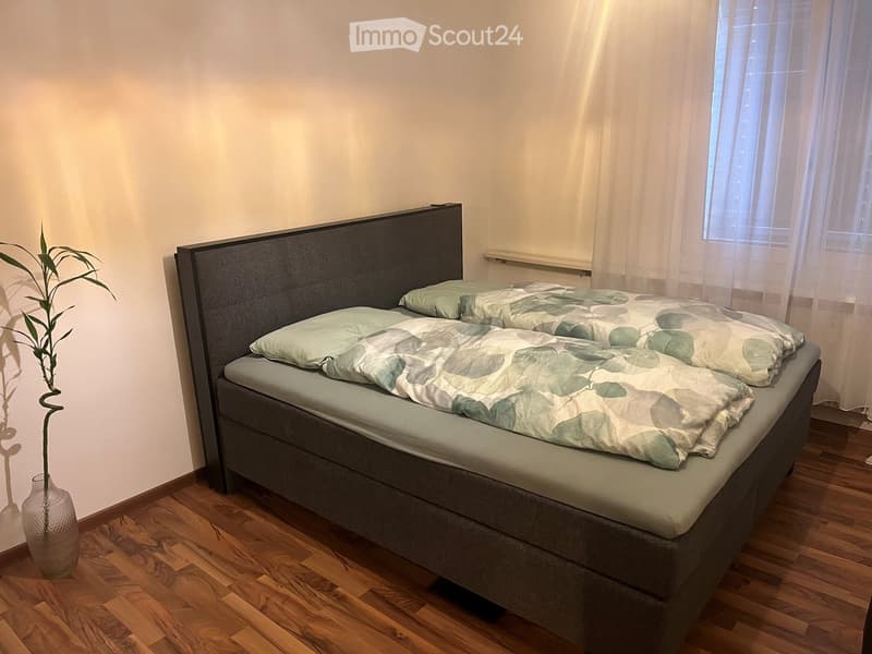 A modern bedroom with a large bed, grey headboard, green and white bedding, a vase with a leafy plant, and wooden flooring.