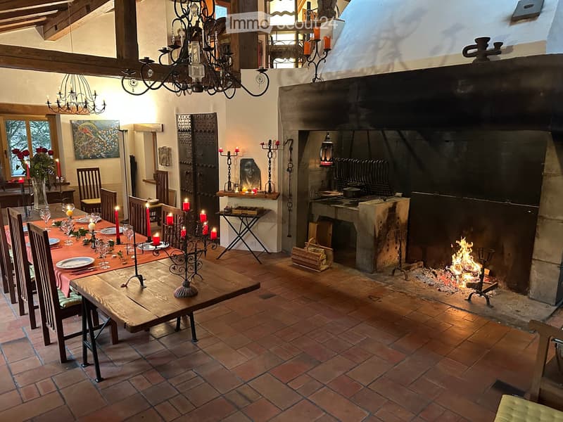 Cozy dining area with a large wooden dining table, chairs, candelabras, and a fireplace with a fire burning. The room features exposed wooden beams, a chandelier, and a painting on the wall.