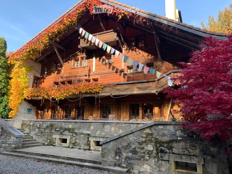 Traditional wooden house with a stone base, multiple windows, and colorful banners hanging from the roofline. The house is surrounded by autumn foliage and a stone staircase leads up to the entrance.