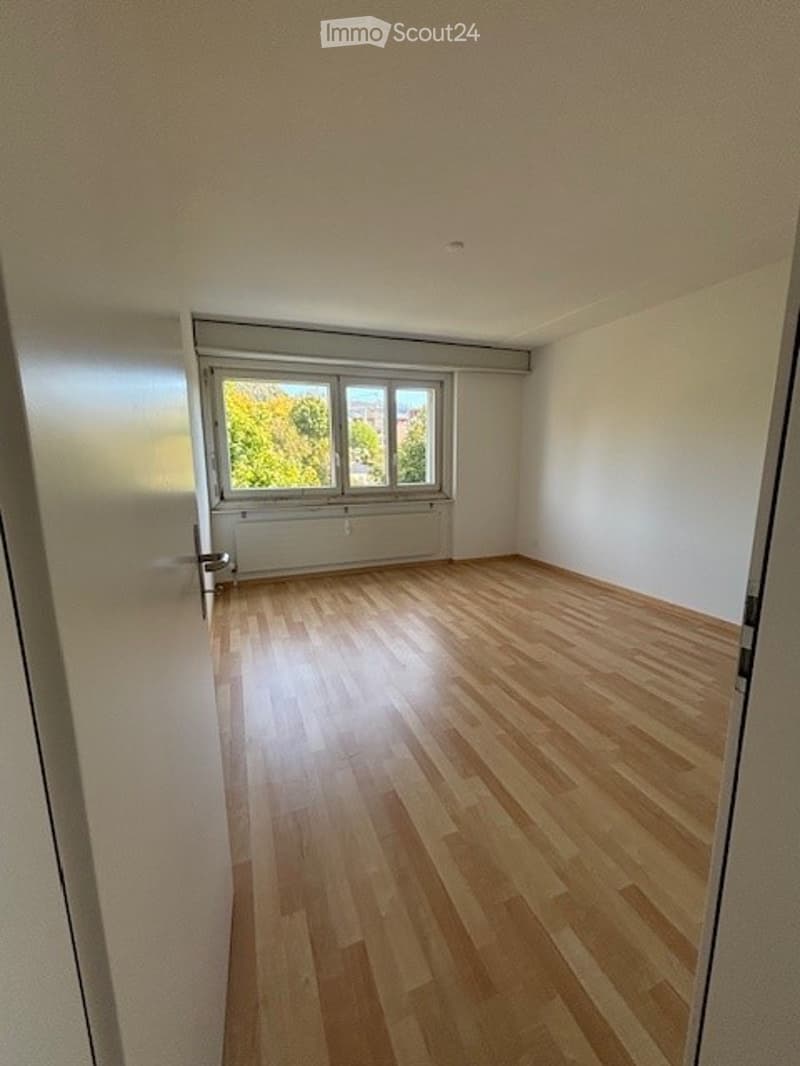 Empty room with wooden flooring, white walls, and a large window with a view of greenery outside.