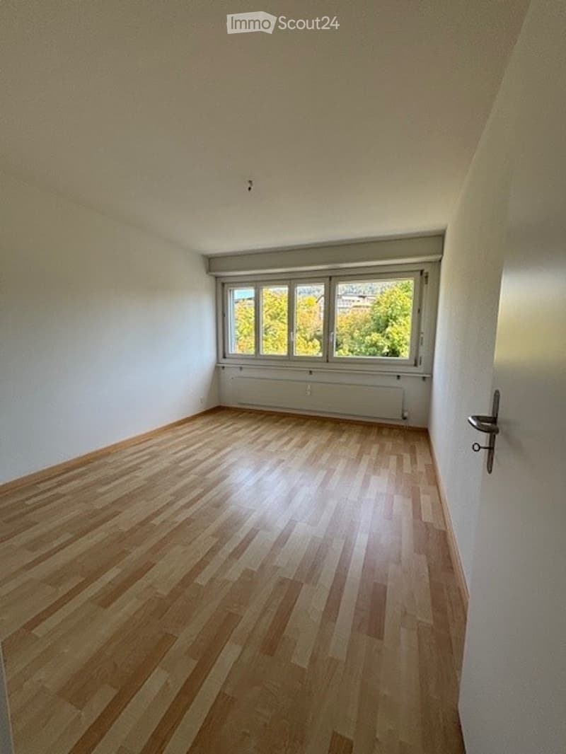 Empty room with wooden flooring, white walls, and large windows with a view of greenery outside.