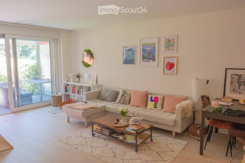Bright living room with a large sectional sofa, coffee table, bookshelf, and a view to a balcony. The room features a mix of modern and cozy decor, including colorful cushions, artwork, and a small dining area.