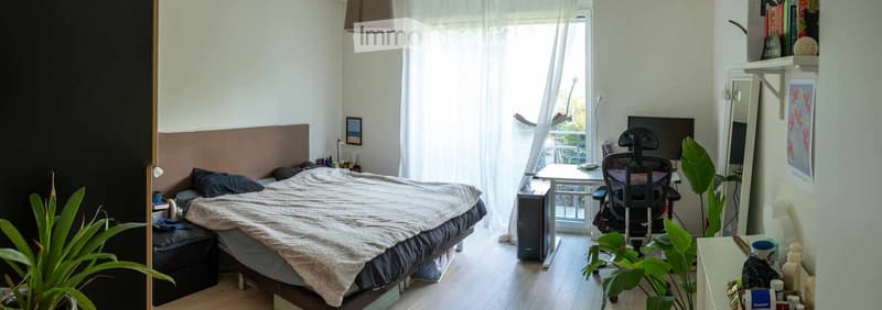 Cozy bedroom with a bed, desk, chair, mirror, and plants. Large window with sheer curtains offers a view of a balcony.