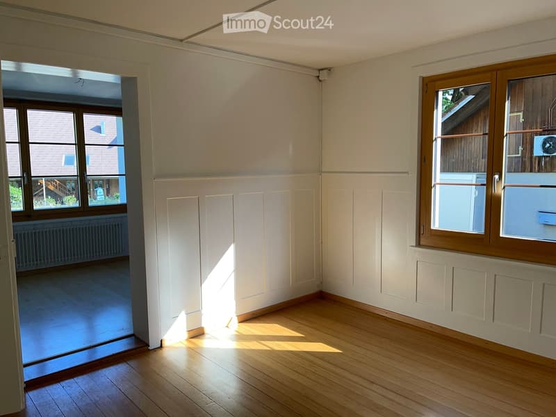 Empty room with wooden flooring, white walls, and large windows allowing natural light to enter. The room features a radiator and a sliding glass door leading to another space.