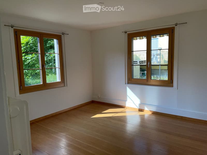 Empty room with wooden flooring, white walls, and large windows with wooden frames. Sunlight streams in, casting shadows on the floor.