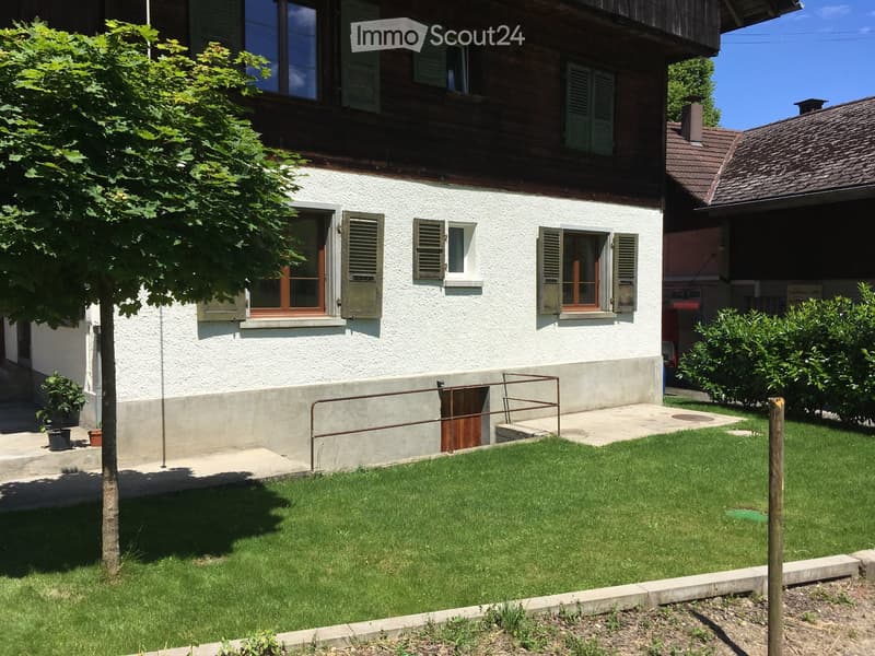 Two-story house with white walls, wooden shutters, and a raised ground floor entrance. The house features a well-maintained lawn and small garden area.