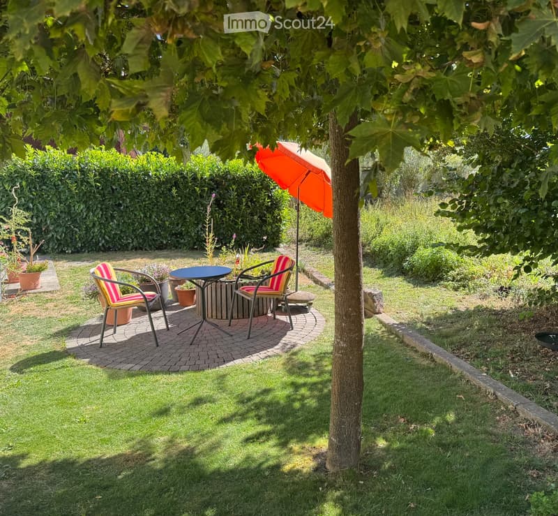 Garden with a round table, two striped chairs, and an orange umbrella under a tree, surrounded by greenery and a hedge.