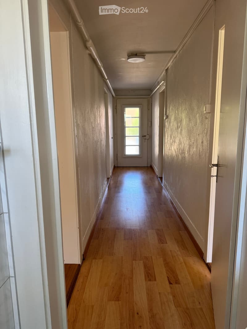 Empty hallway with wooden flooring, white walls, and a single window at the end.
