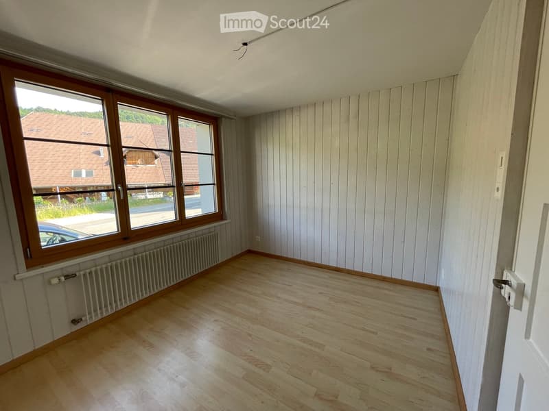 Empty room with wooden flooring, white paneled walls, large windows with wooden frames, and a radiator under the windows.