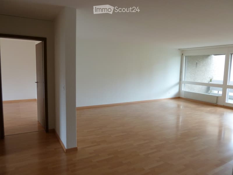 Empty room with wooden flooring, white walls, and large windows allowing natural light. A door is visible on the left side.