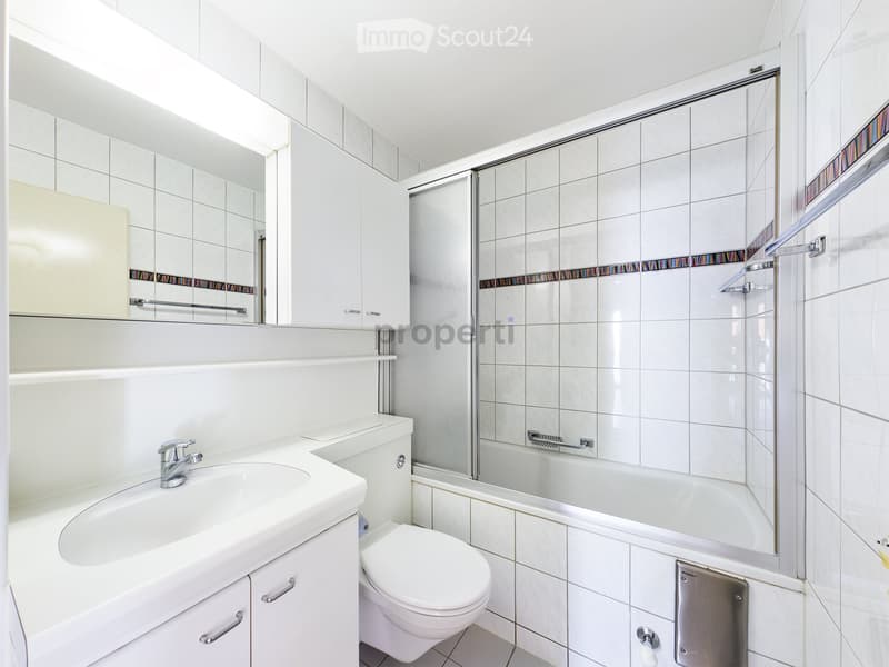 Modern bathroom with white tiles, a sink with a faucet, a mirror, a toilet, a bathtub, and a glass-enclosed shower area.
