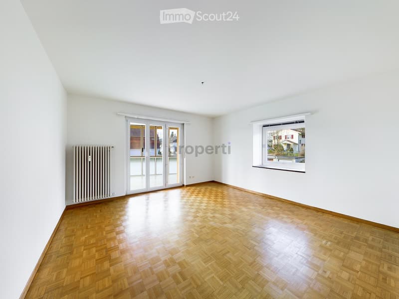 Empty room with wooden parquet flooring, white walls, large window, sliding glass door, and radiator.