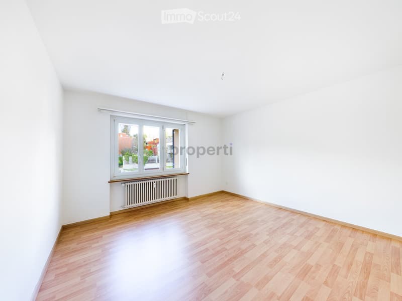 Empty room with light wooden flooring, white walls, and a large window with a radiator below it.