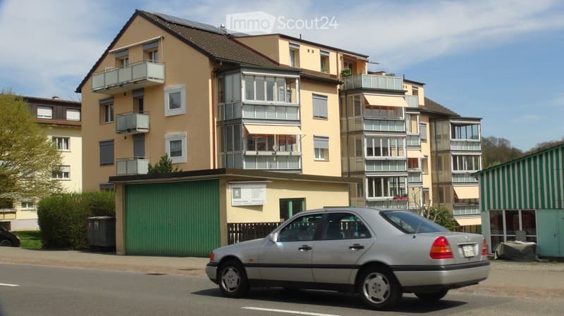 Multi-story residential building with balconies, solar panels on the roof, and a garage. A silver car is parked in front of the building.