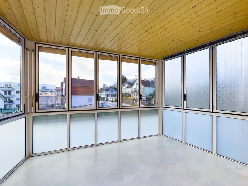 Bright, empty room with large windows and a wooden ceiling, featuring a light-colored tiled floor.
