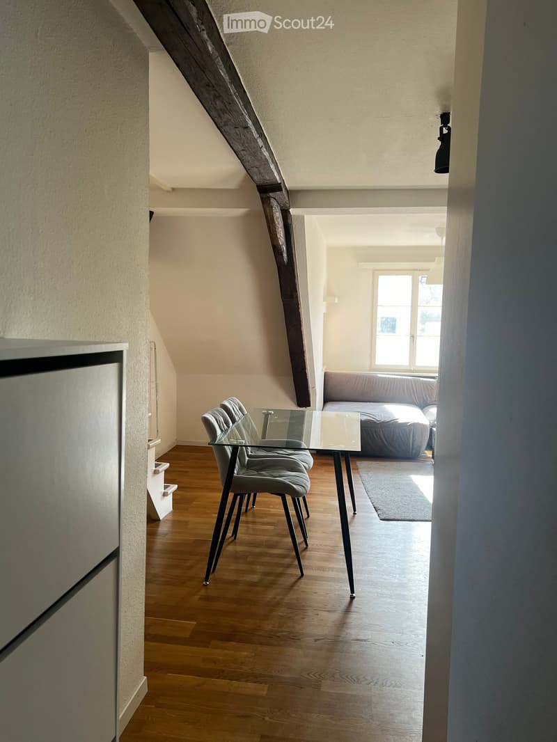 Bright, airy room with wooden flooring, a glass dining table with four chairs, and a grey sofa against the wall. Natural light streams in from a large window.