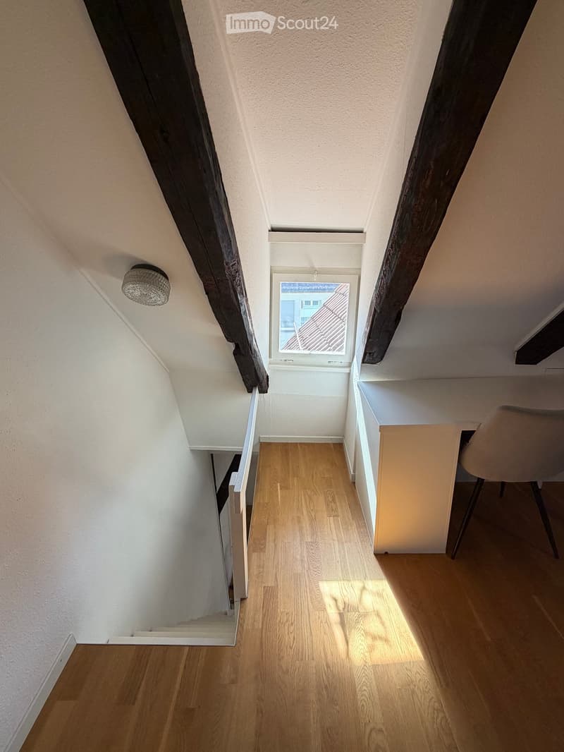 Bright, narrow hallway with wooden flooring, a small desk, and a chair, leading to a window with a view of a roof.