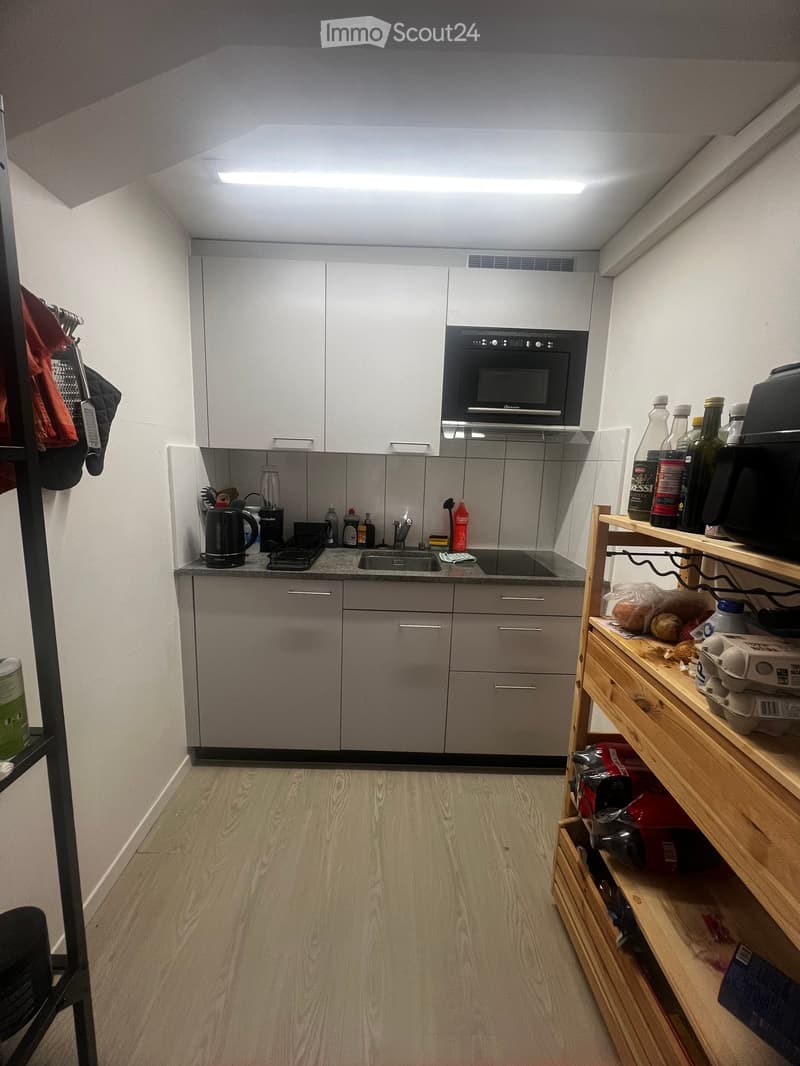 Modern kitchen with white cabinets, stainless steel appliances, and wooden shelves. The space includes a sink, oven, microwave, and various kitchen items on the counter and shelves.