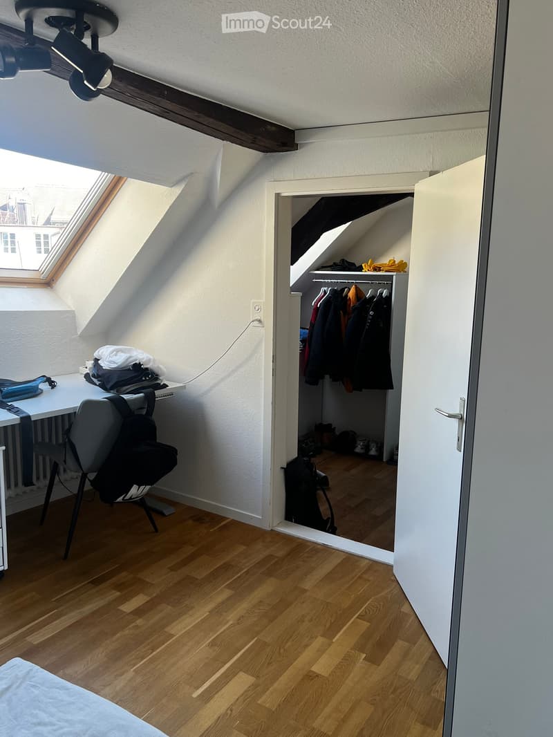 A small attic room with wooden flooring, a desk with a chair, a backpack, and an open closet with clothes and shoes.