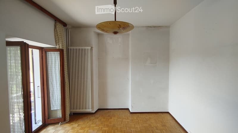Empty room with wooden flooring, white walls, a radiator, and a large window with curtains. The room has a simple ceiling light fixture.