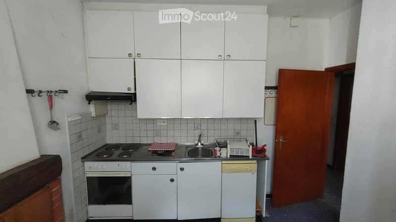 A compact kitchen with white cabinets, a four-burner electric stove, a sink, an oven, and a dishwasher. The walls are tiled, and there is a small countertop with a few items, including a cup and a spoon.