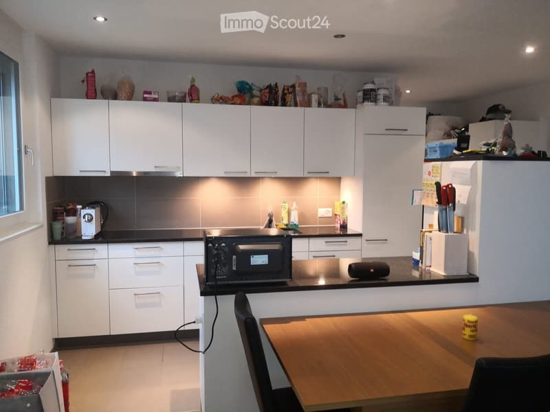 White kitchen cabinets, black countertops, island with seating, black microwave, coffee machine, toaster, refrigerator, utensils, storage boxes, various kitchen items