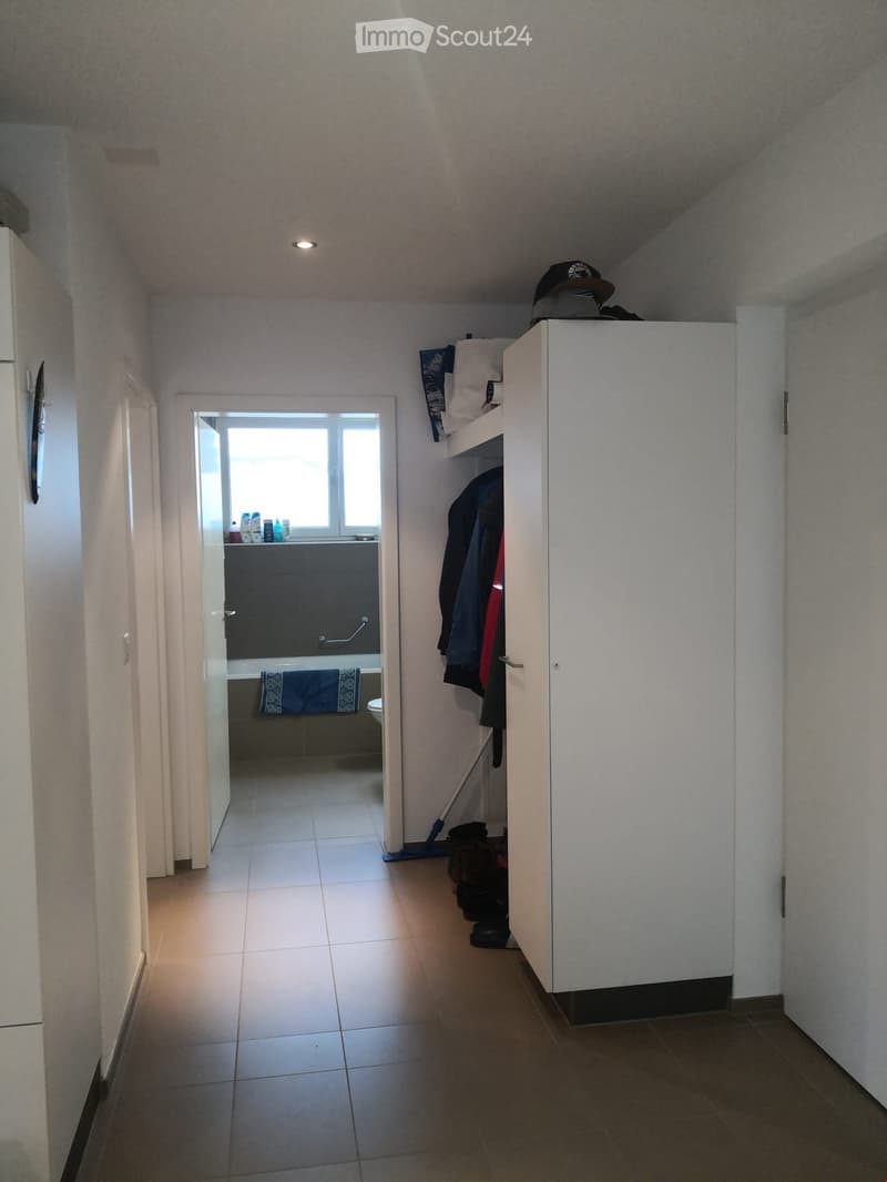 A corridor with tiled floor and white walls leading to a bathroom. In the hallway, there is a white wardrobe with clothes inside and a door on the left side. The bathroom has a bathtub with a towel, a toilet, and shelves with cleaning products. In the dis