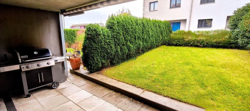 Well-maintained garden with neatly trimmed hedges, a small lawn, and a modern barbecue grill on a tiled patio.