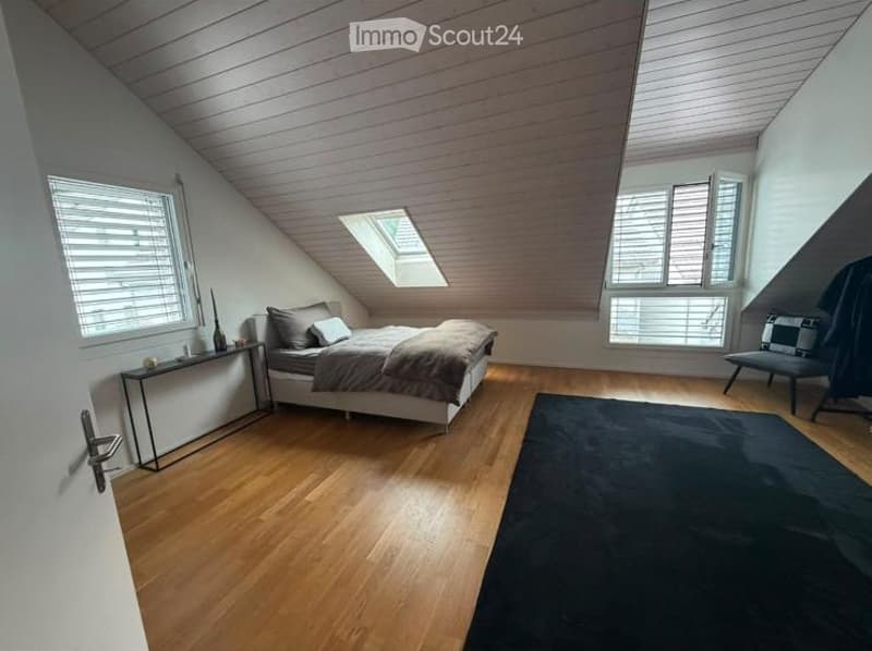 Bright, modern bedroom with wooden floors, slanted ceilings, large windows, and a skylight. Features a bed with pillows and a duvet, a nightstand with a vase, a black rug, and a chair with a patterned cushion.