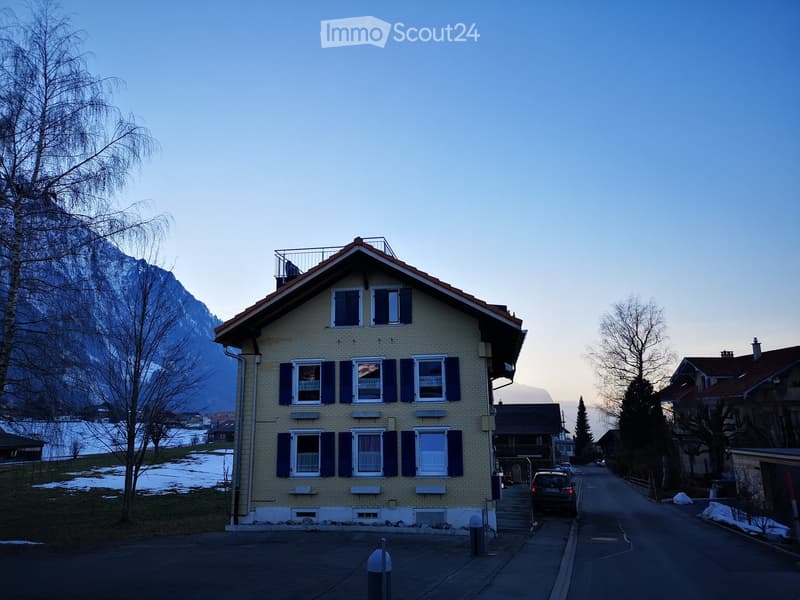 Two-story yellow house with blue shutters, white-framed windows, and a small balcony on the roof. Snow-covered ground and mountains in the background.