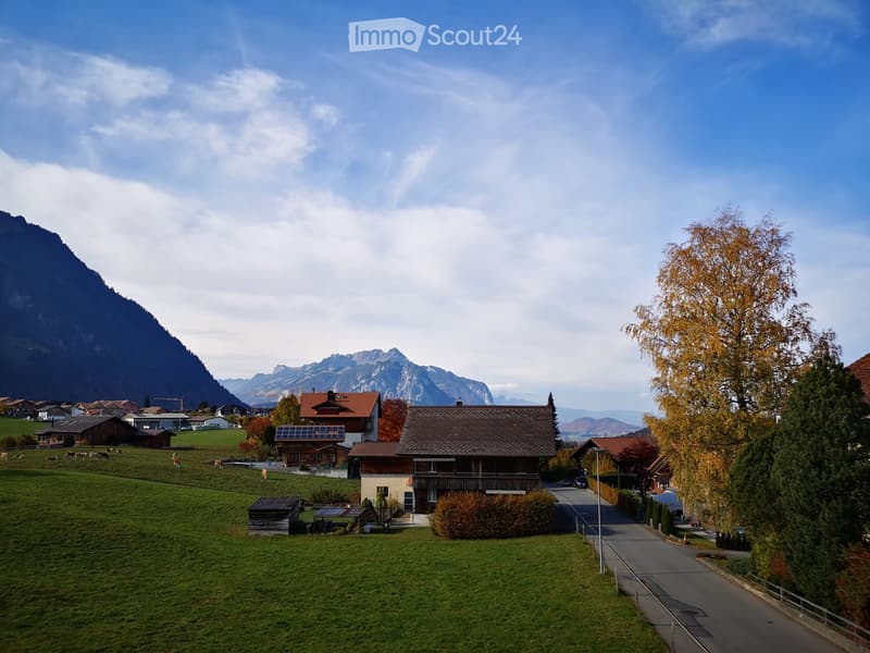 Scenic rural village with traditional houses, green fields, grazing cows, and mountain backdrop.