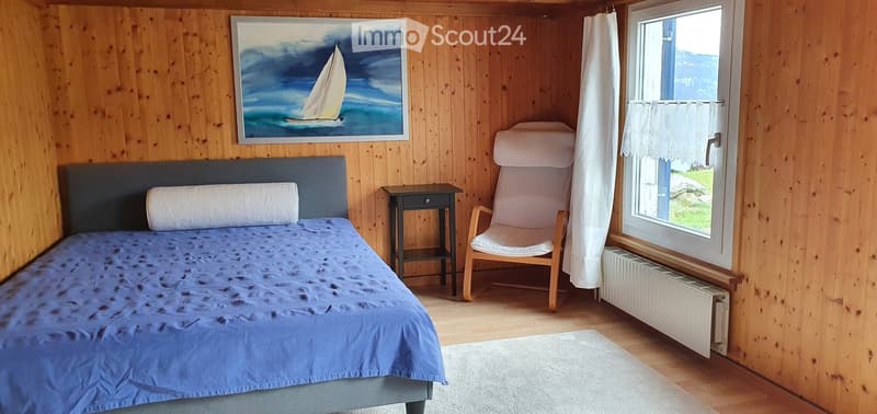 Cozy wooden bedroom with a double bed, nightstand, armchair, and a painting of a sailboat on the wall.