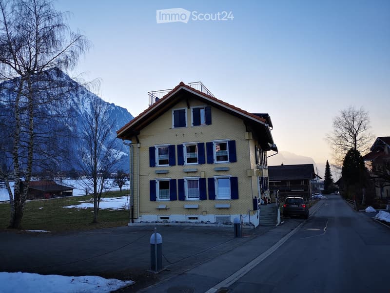 Two-story house with yellow brick facade, blue shutters, and white trim. Features multiple windows, a small balcony on the roof, and is situated in a snowy landscape with mountains in the background.