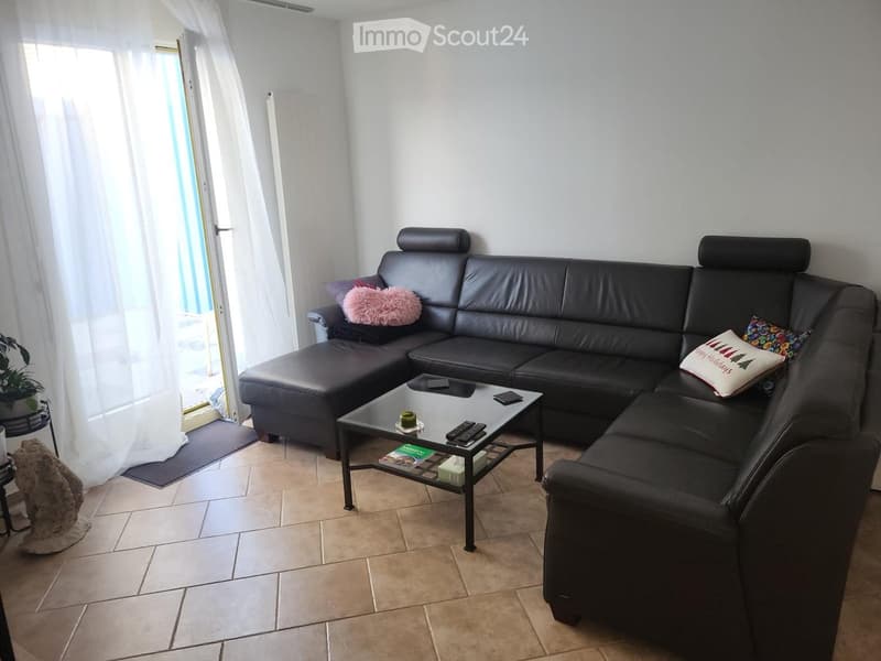 Modern living room with a black leather sectional sofa, a glass coffee table, and a small plant in the corner. The room features tiled flooring and large windows with white curtains.