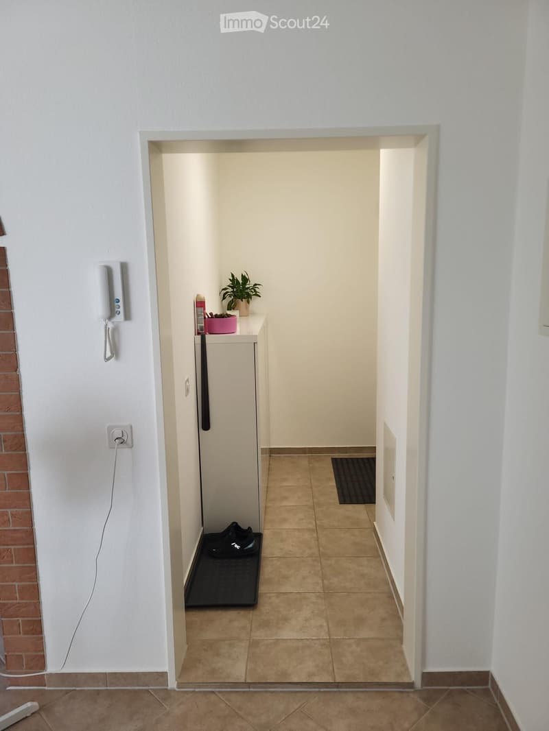A narrow hallway with tiled flooring, white walls, and a small shelf with a plant and other items. A black bag is placed on the floor near the shelf.