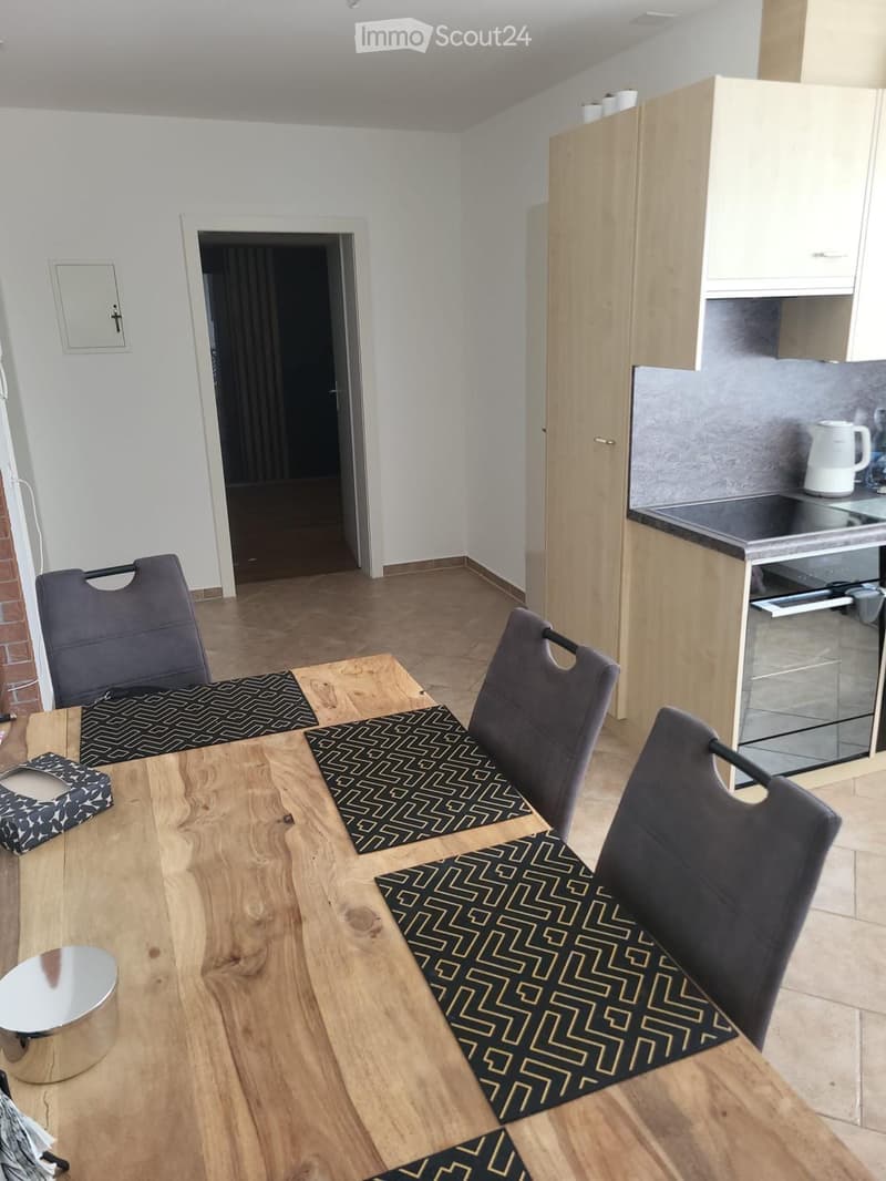 Modern dining area with a wooden dining table, four dark grey chairs, and a small kitchen area with cabinets and an oven. The floor is tiled, and the walls are white.