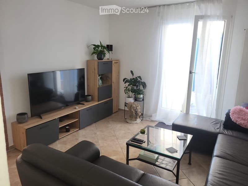 Modern living room with a large TV, a black leather sofa, a glass coffee table, and a wooden TV stand. There are plants and decorative items on the TV stand and a small table.