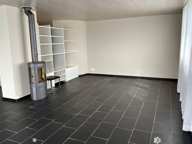 Empty room with dark tiled floor, white walls, and a modern wood-burning stove with a chimney pipe. A small black bench is placed next to the stove, and a white shelving unit is visible in the background.