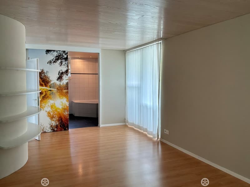 Empty hallway with wooden flooring, white walls, and a large window with white curtains. A doorway leads to a tiled bathroom with a bathtub.