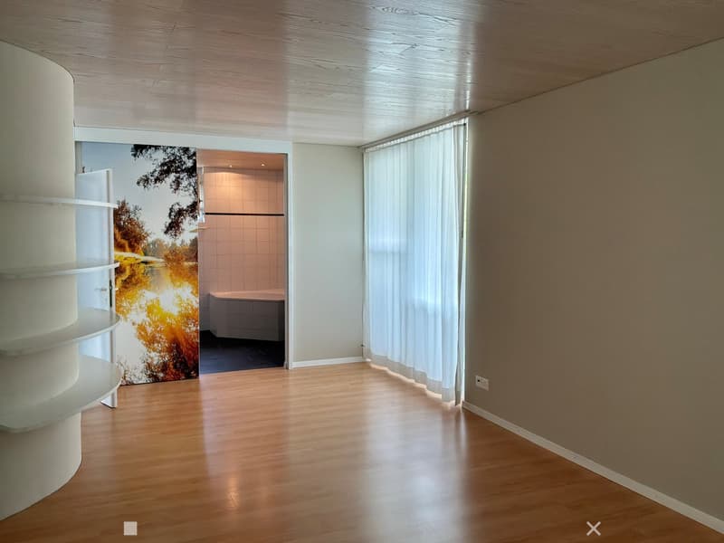 Empty hallway with wooden flooring, white walls, and a large window with white curtains. A doorway leads to a tiled bathroom with a bathtub.