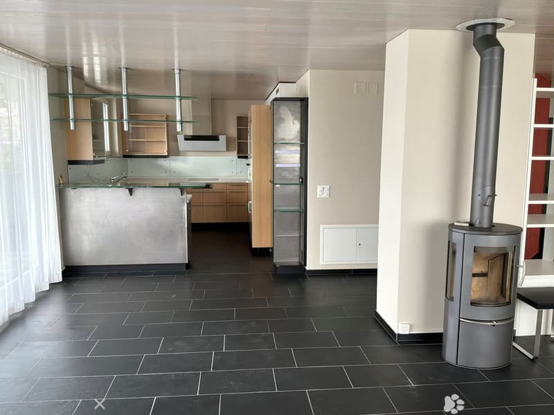 Modern kitchen with dark tiled flooring, light-colored cabinets, glass shelves, and a central island. A wood-burning stove is positioned near the wall with a chimney pipe extending to the ceiling.