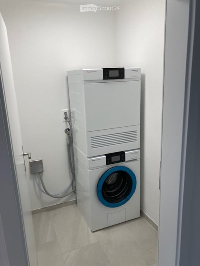 A compact bathroom with a stacked washing machine and dryer unit, white tiled floor, and white walls.