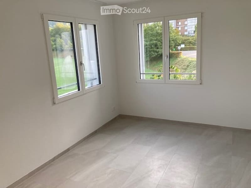 Empty room with white walls, tiled floor, and large windows offering a view of greenery outside.